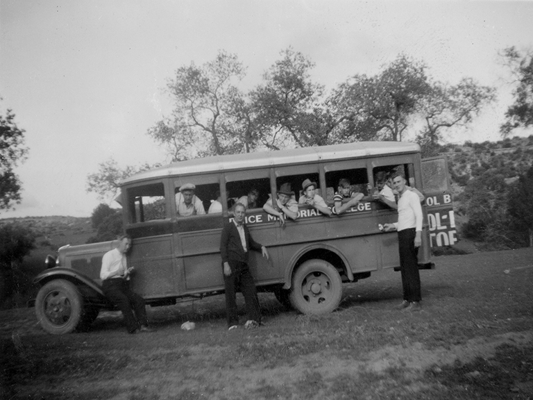 Three men stand outside a small bus marked “Price Memorial College”, with about a dozen boys, mostly all in hats, leaning out the open windows.  The bus is parked in an open field.